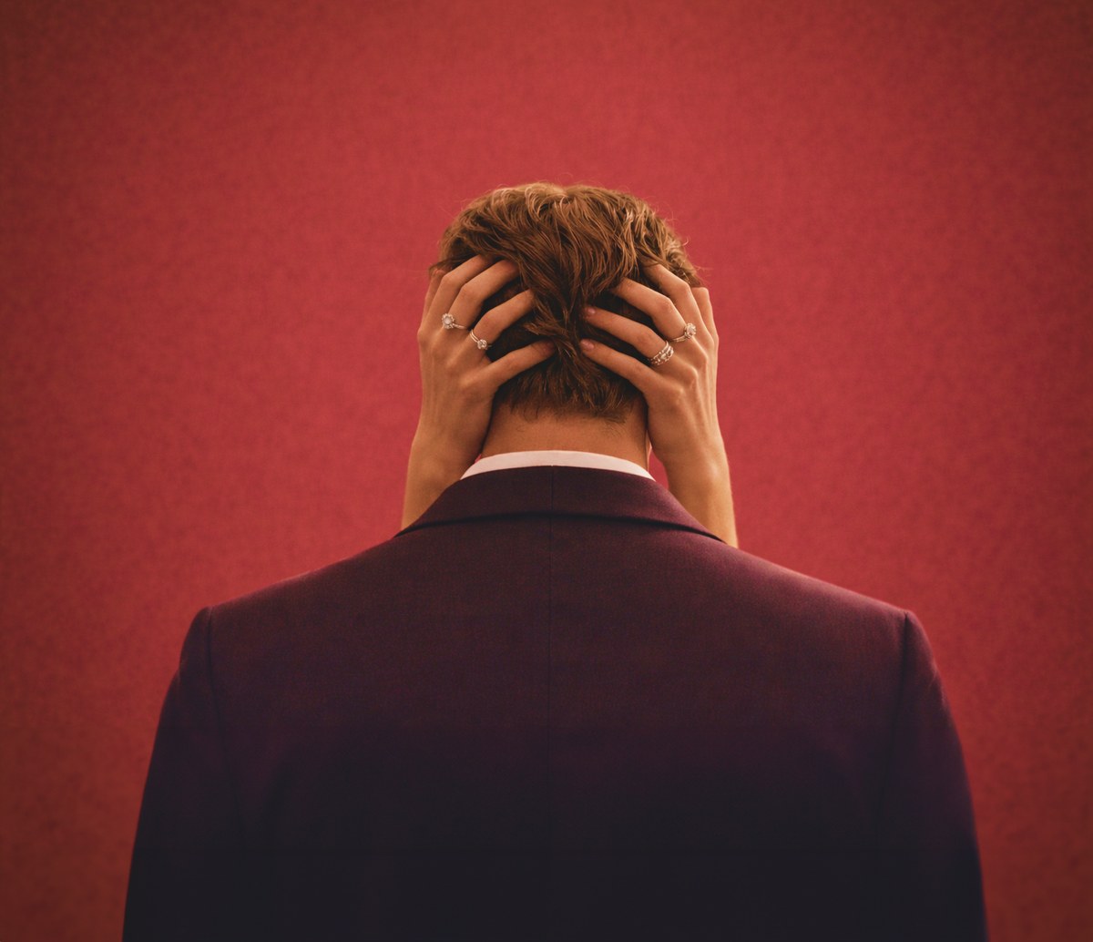 The Husband gripping his own hair with both hands, seen from behind against a red wall.
